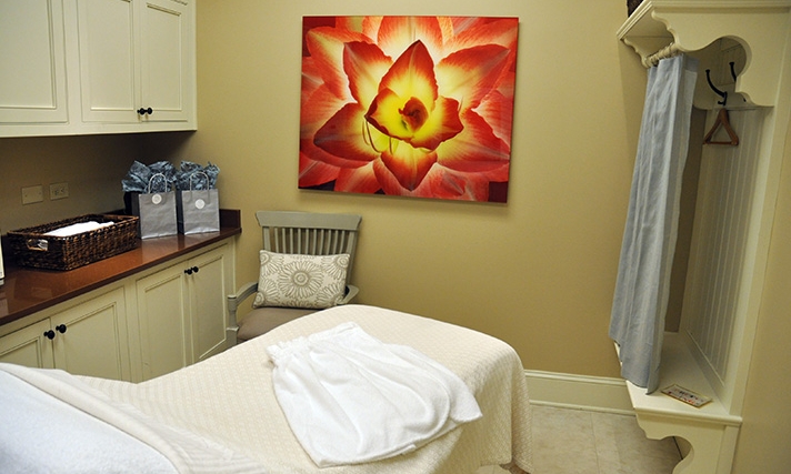 A spa room with a massage table, a folded towel, a chair with a pillow, a large flower painting on the wall, and a counter with gift bags and a wicker basket.